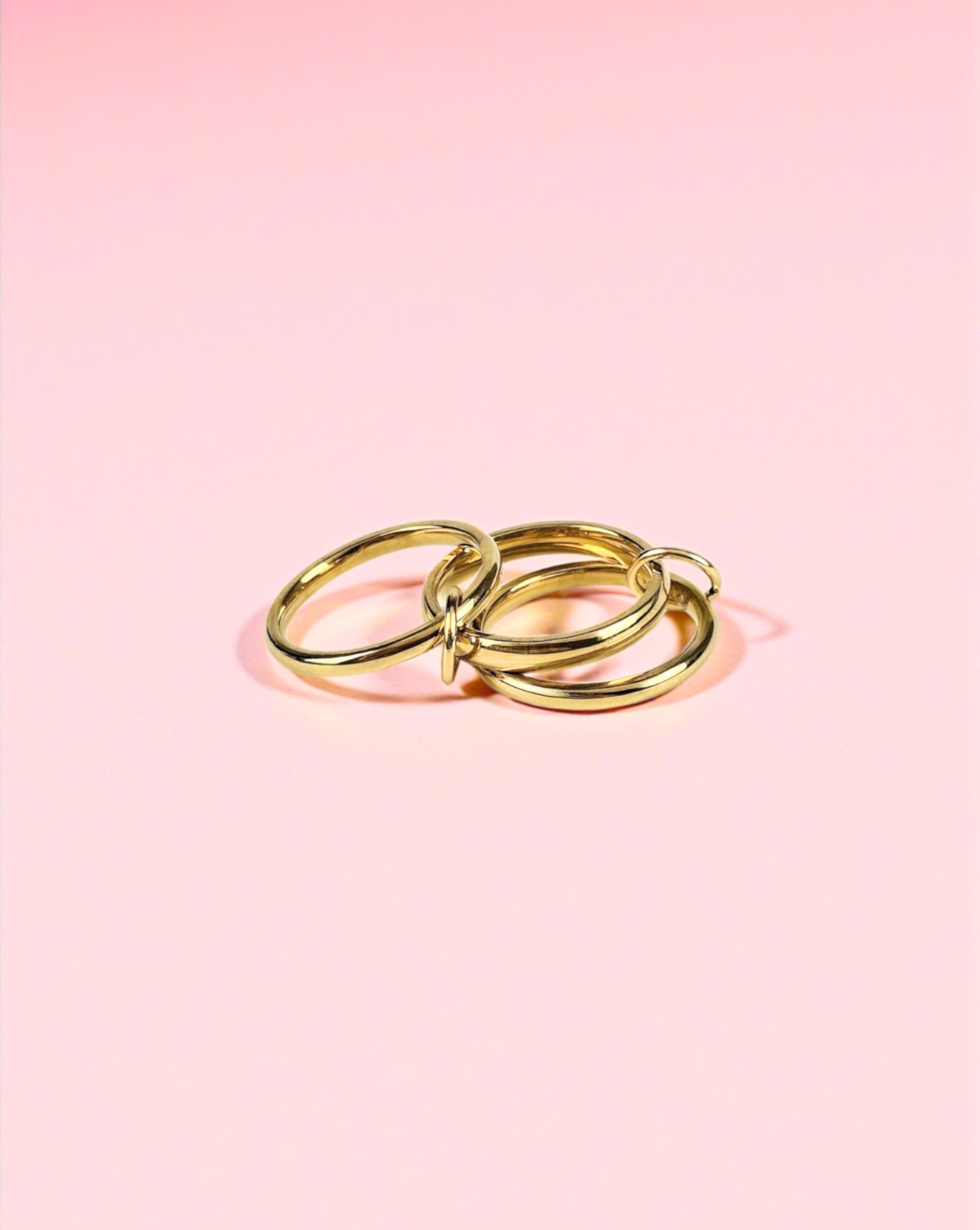 Prstan TRIPPLE BANDS Gold iz nerjavečega jekla s tremi povezanimi obročki, fotografiran na rožnatem ozadju – Trgovina Patrizia Ljubljana.